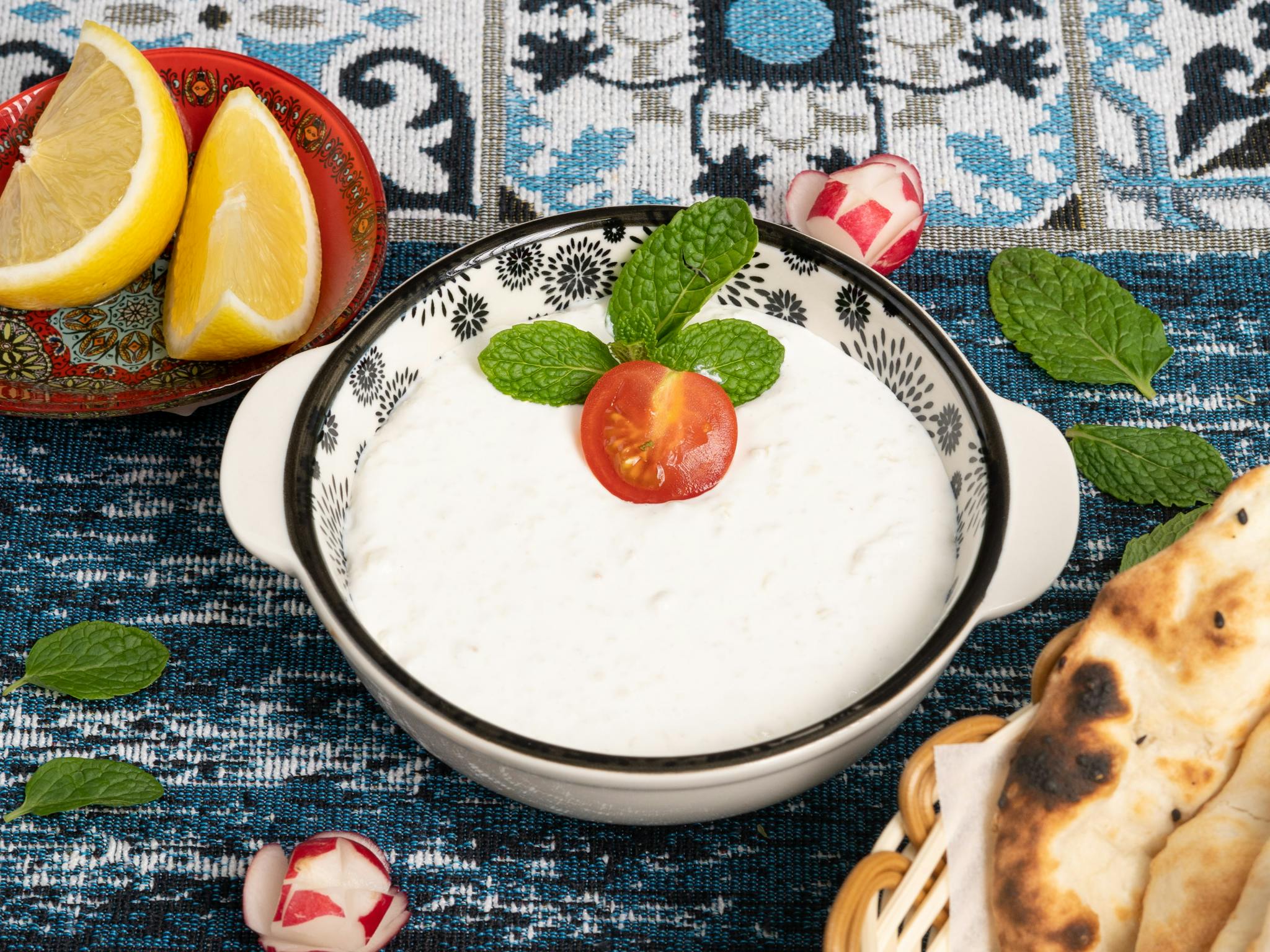 Delicious bowl of creamy yogurt garnished with mint leaves and a cherry tomato, surrounded by fresh bread and lemon slices.